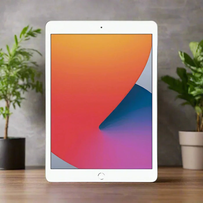 A certified refurbished Apple iPad 8 Cellular Silver 32GB, 10.1" display, stands upright on a wooden surface showing a colorful screen. Two potted plants are in the background against a gray wall. Brand: Apple.