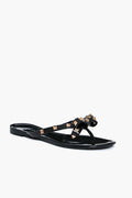 The Anina black thong sandals feature a flat sole, bow detail, and gold pyramid studs on the straps. These jelly bowtie sandals add a trendy, stylish touch to any outfit.