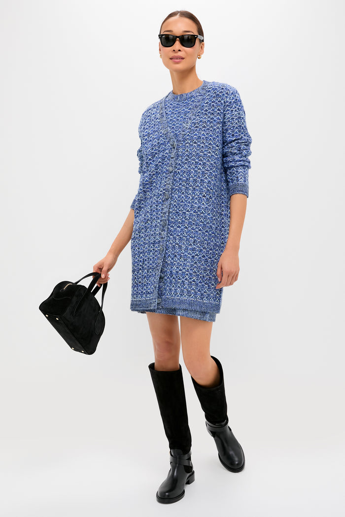 A woman showcases effortless chic in Tuckernuck sunglasses, a blue patterned knit dress, black knee-high boots, and a black handbag, posed against a clean white background.