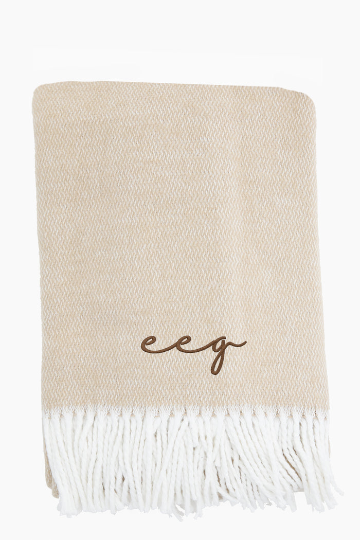 The Matouk beige brushed cotton throw features white fringed edges and brown "eeg" embroidered initials for a touch of luxury.