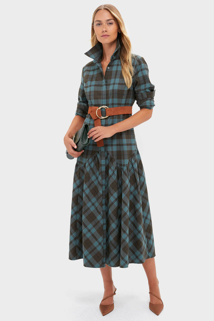 A woman in a Tuckernuck blue and brown plaid dropped-waist dress with a brown belt, carrying a dark green clutch and wearing brown pointed-toe shoes.