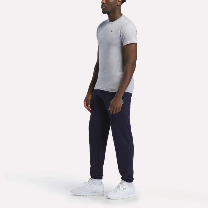 A man stands against a plain white background wearing a relaxed fit Reebok Men's Small Logo T-Shirt in grey, dark blue jogger pants, and white sneakers.