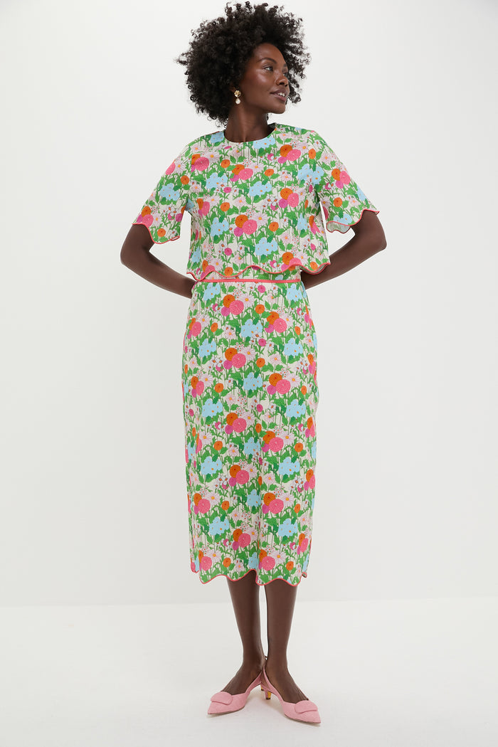 A woman stands against a plain background in a Hyacinth House Multi Green Begonia Floral midi skirt and matching pink, green, and white top paired with light pink pointed shoes.