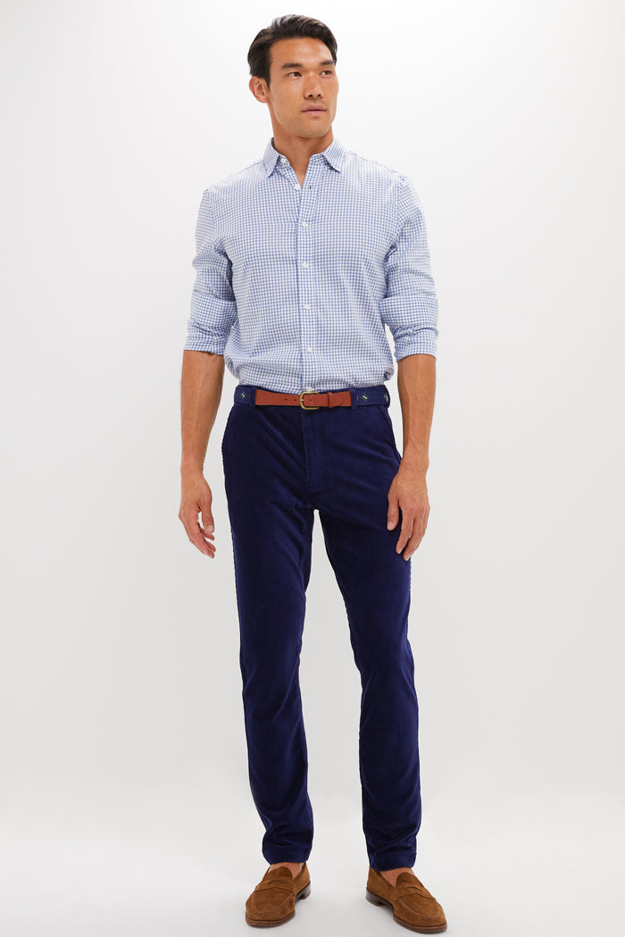A man wears a light blue checkered shirt, blue REDVANLY performance corduroy trousers, a brown belt, and brown loafers against a plain white background.