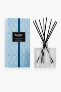 A NEST New York Ocean Mist & Sea Salt Reed Diffuser in a rectangular glass bottle with black reeds stands by its patterned light blue box, offering a cruelty-free home fragrance.