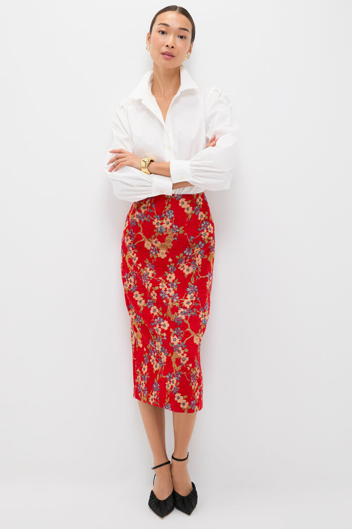 A person poses against a white background wearing a Tuckernuck white collared shirt, a chic floral jacquard skirt, black pointed shoes, and a gold watch, arms crossed.
