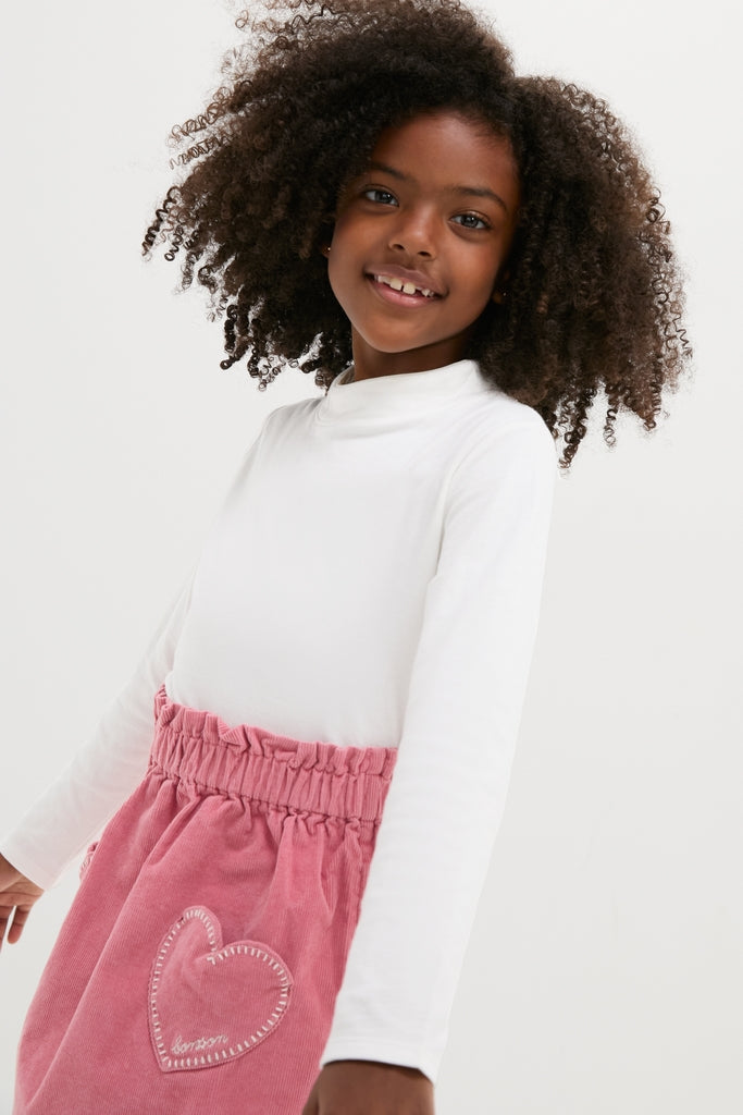 Young girl in a White Frank T-Neck and pink skirt, showcasing Oso & Me's classic, effortless design. Perfect for fun, high-end, versatile styling.