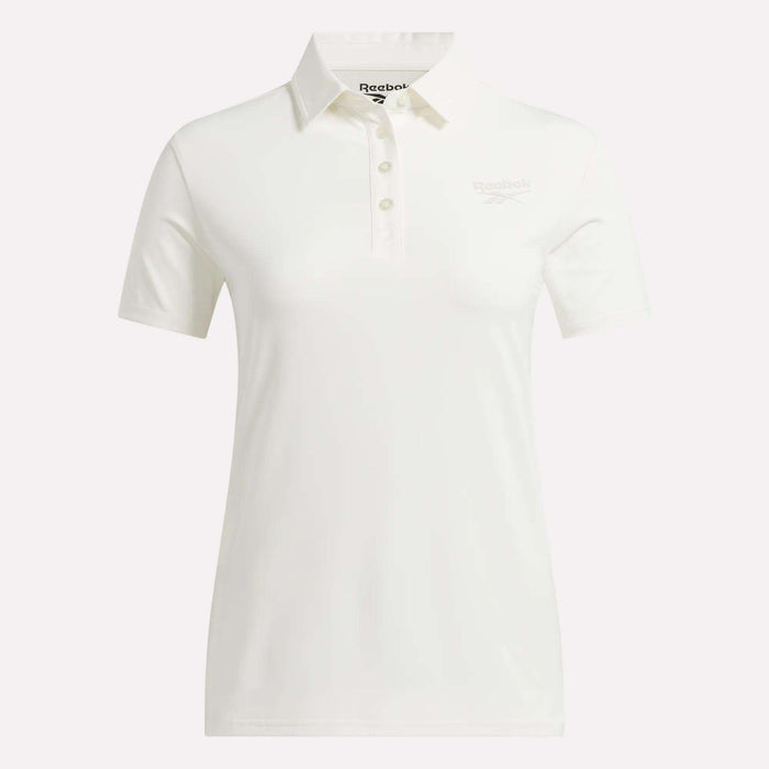The Reebok Women's Solid Core Polo Shirt is a white, short-sleeve golf top featuring a classic collar and button placket. Made from performance jersey for comfort and breathability, it displays a subtle Reebok logo on the left chest. The shirt is shown against a plain white background.