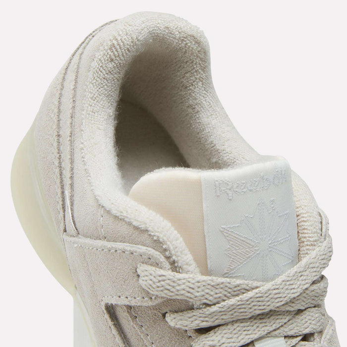 Close-up of Reebok Women's Workout Plus Shoes in light beige, featuring suede texture, thick laces, and a partially visible tongue logo. Plush inner lining adds comfort, capturing an '80s fitness look ideal for women's workout footwear.